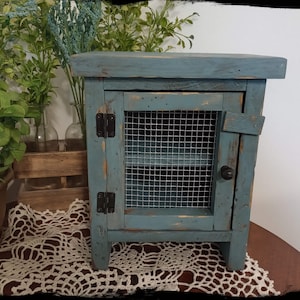 May include: A small, blue wooden cabinet with a wire mesh door and a single shelf. The cabinet has a distressed paint finish and black hinges.