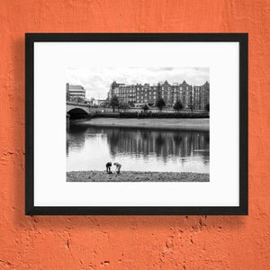 May include: Black and white framed photograph of a river scene. Two figures are bent over on the riverbank. A bridge and buildings are in the background, with reflections in the water. The photograph is in a black frame.