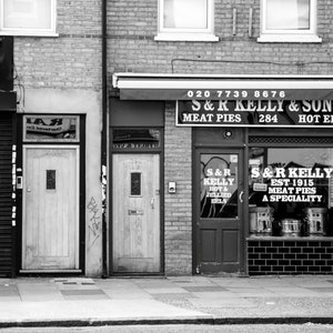 May include: Black and white photo of a storefront with a sign that reads "S&R Kelly & Sons Meat Pies 284 Hot Eels". The storefront has two doors and a window with a sign that reads "S&R Kelly Est 1915 Meat Pies A Speciality".