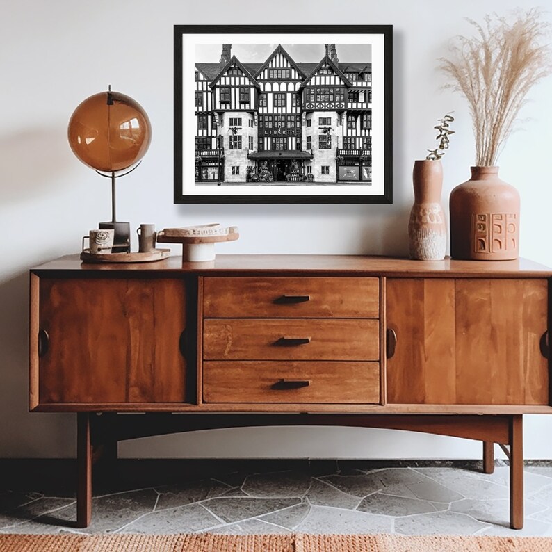 May include: Black and white photograph of a large, ornate building with a storefront. The building has a multi-level facade with multiple windows and a large entrance. The building is framed by a black picture frame and is displayed on a wooden dresser with a globe lamp and decorative vases.