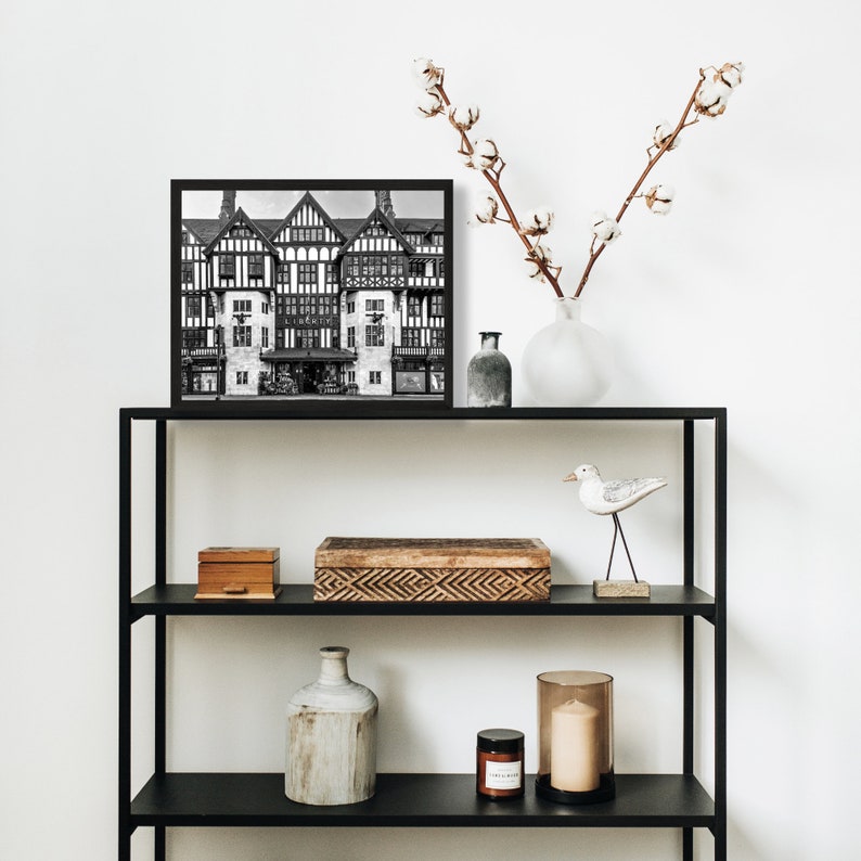 May include: Black and white photograph of a building with a black frame, displayed on a black metal shelf with other decorative items, including a wooden box, a ceramic vase, a glass candle holder, and a white bird figurine.