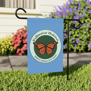 May include: A blue garden flag with a circular design featuring an orange butterfly and the words "Pollinator Habitat" and "Native plants live here!". The flag is displayed on a black metal stand in a garden setting.