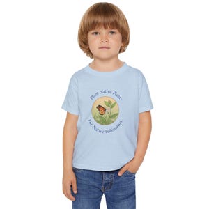 May include: A light blue t-shirt featuring a circular graphic with a monarch butterfly, plants, and the text "Plant Native Plants For Native Pollinators." The shirt is worn by a young child, and is paired with blue jeans.