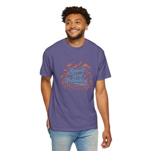 Puede incluir: Una camiseta morada con un diseño gráfico de flores y el texto "Support Local Pollinators" en azul y naranja.