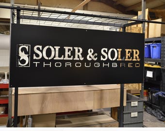 Op de afbeelding: Een rechthoekig zwart metalen bord met de uitgesneden tekst SOLER & SOLER en THOROUGHBRED in een stencil-stijl lettertype. Een vierkant logo met een gestileerde letter S en een ster bevindt zich links van de hoofdtekst op het bord.