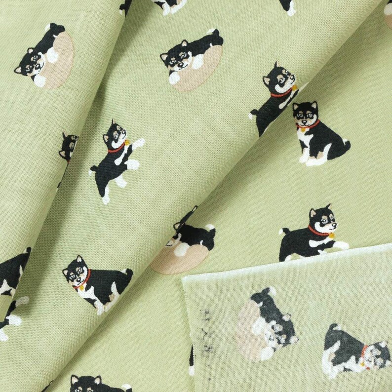 May include: Green fabric with a repeating pattern of black and white Shiba Inu dogs. Some dogs are lying down, while others are standing and looking to the right. The fabric is folded in half, showing the pattern on both sides.