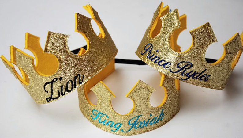 May include: Three gold glitter crowns with black elastic bands. The crowns are decorated with the names "Lion", "King Josiah", and "Prince Ryder" in different colored embroidery.