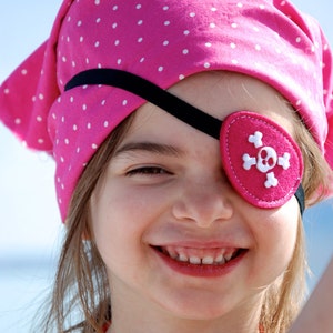 May include: A bright pink bandana featuring a white polka dot pattern, paired with a matching pink felt eye patch that displays a white stitched skull and crossbones design, secured by a thin black elastic strap across the forehead.