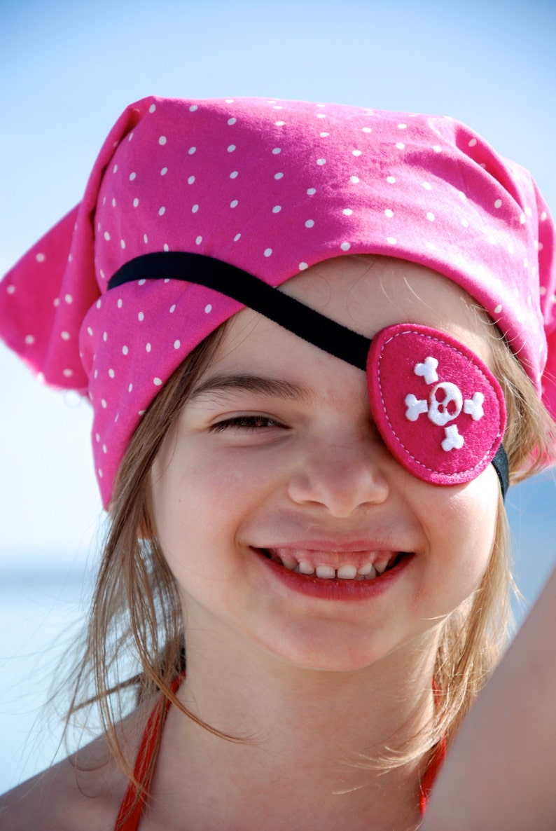 May include: A bright pink bandana featuring a white polka dot pattern, paired with a matching pink felt eye patch that displays a white stitched skull and crossbones design, secured by a thin black elastic strap across the forehead.