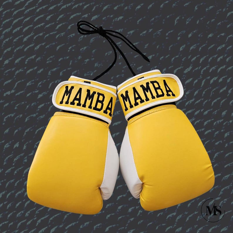 May include: Pair of yellow and white boxing gloves with the word "MAMBA" in black lettering on the wristbands. The gloves are hanging by a black string against a textured gray background.
