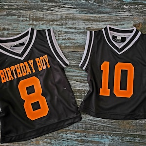 May include: Two black basketball jerseys with white trim. One jersey has "Birthday Boy" and the number "8" in orange. The other jersey has the number "10" in orange.