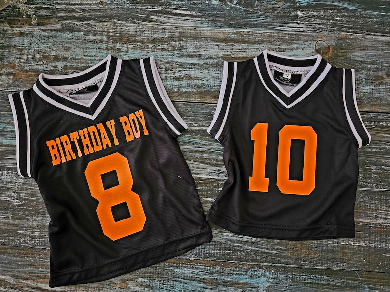 May include: Two black basketball jerseys with white trim. One jersey has "Birthday Boy" and the number "8" in orange. The other jersey has the number "10" in orange.