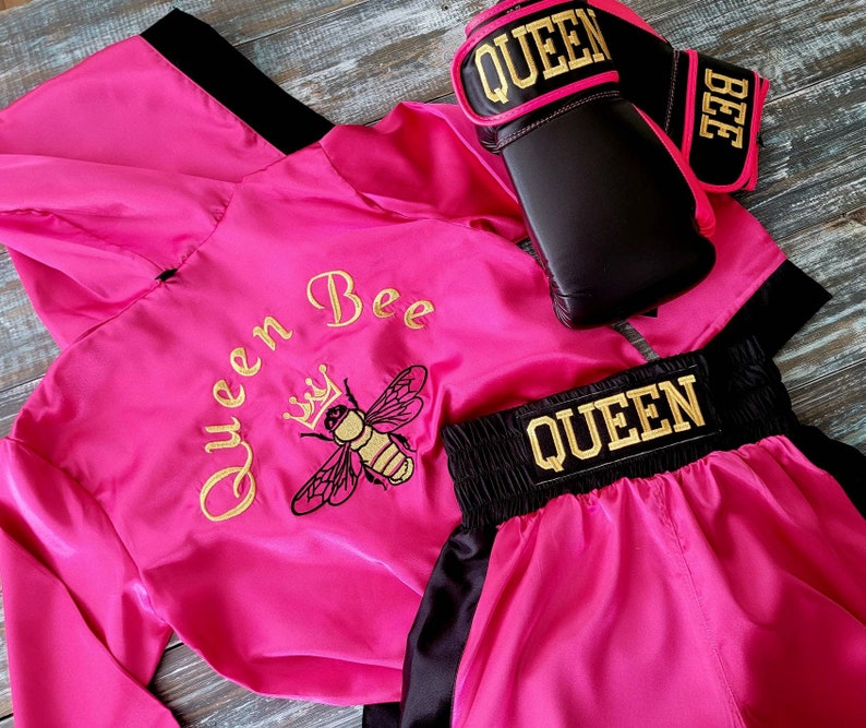 May include: A hot pink boxing outfit set with black accents. The set includes a hooded robe, boxing gloves, and shorts. The robe features "Queen Bee" and a bee graphic embroidered in gold. The gloves and shorts have "QUEEN" in gold lettering.
