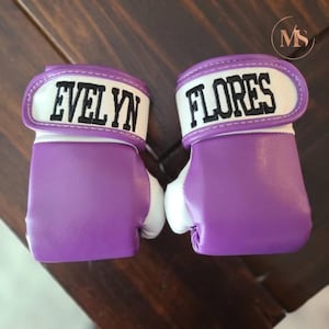 May include: Pair of purple and white boxing gloves. The gloves have the names "EVELYN" and "FLORES" embroidered in black on the white wrist straps. The gloves are made of a smooth material and are designed for boxing.
