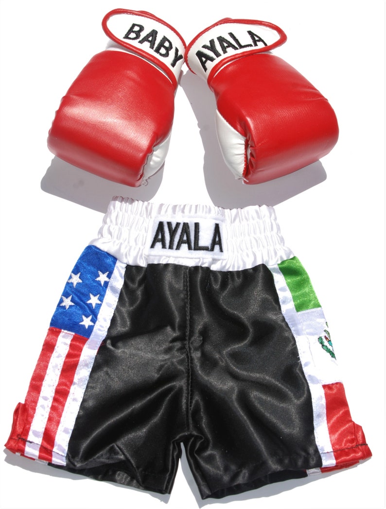 May include: Red and white boxing gloves with the name "BABY AYALA" on them, and black boxing shorts with the name "AYALA" on the waistband. The shorts have an American flag on one side and a Mexican flag on the other.