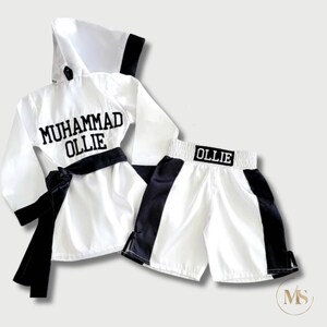 May include: A white boxing robe and shorts set. The robe has a hood, black trim, and a black belt. The name "MUHAMMAD OLLIE" is embroidered on the robe. The shorts are white with black side panels and the name "OLLIE" on the waistband.