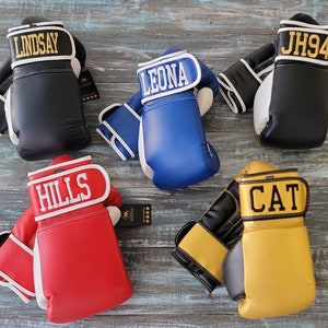 May include: An assortment of personalized boxing gloves in various colors, including black, blue, red, and gold. Each glove features a name or initials in bold lettering on the wrist strap. The gloves are displayed on a weathered wooden surface.