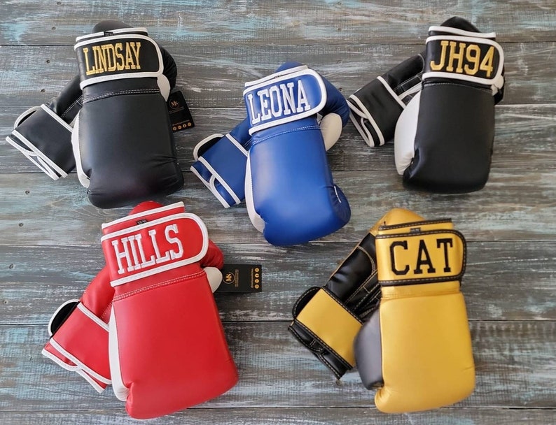 May include: An assortment of personalized boxing gloves in various colors, including black, blue, red, and gold. Each glove features a name or initials in bold lettering on the wrist strap. The gloves are displayed on a weathered wooden surface.