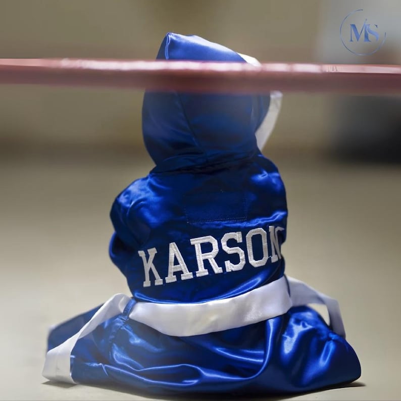 May include: A blue boxing robe with white trim and the name "KARSON" embroidered in white. The robe is made of a shiny, satin-like material and is draped over a boxing ring's ropes.