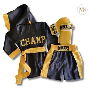 May include: A black and gold boxing outfit set, including a robe, shorts, and gloves. The word "CHAMP" is embroidered in gold on the robe, shorts, and gloves. The robe has a hood and a gold belt.