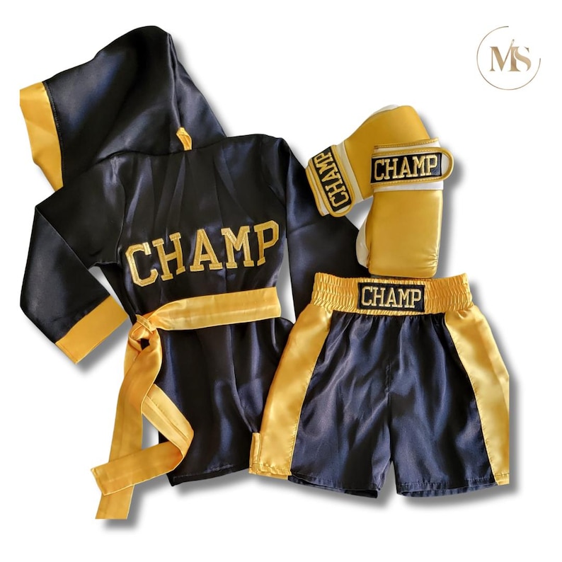 May include: A black and gold boxing outfit set, including a robe, shorts, and gloves. The word "CHAMP" is embroidered in gold on the robe, shorts, and gloves. The robe has a hood and a gold belt.