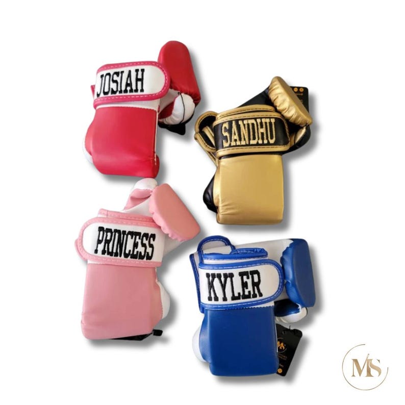 May include: Four personalized boxing glove ornaments in red, gold, pink, and blue. Each glove has a name printed on the wristband: Josiah, Sandhu, Princess, and Kyler. The gloves are set against a white background.