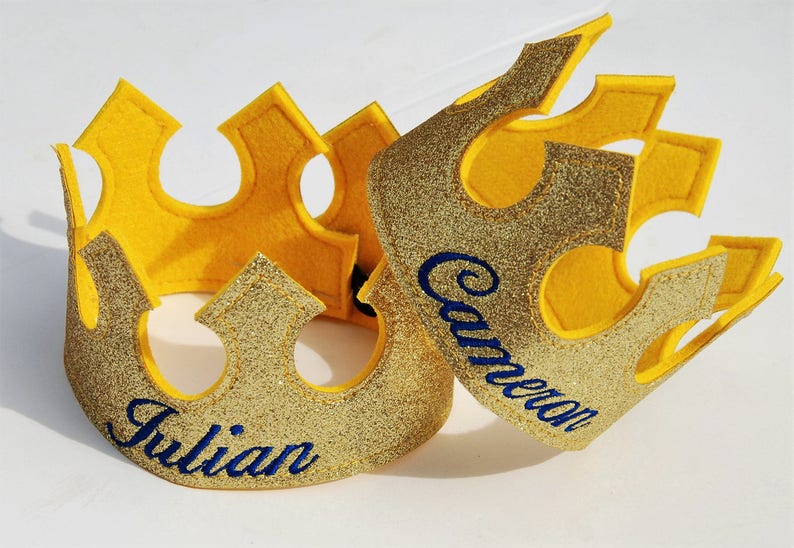 May include: Two gold glitter crowns with yellow felt backing. The crowns are decorated with blue embroidered names, "Julian" and "Cameron".
