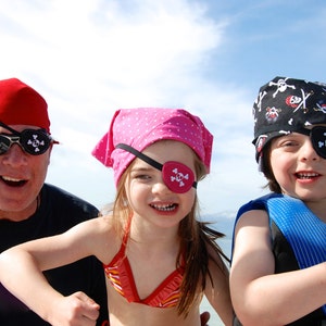 May include: Three people wearing pirate eye patches and bandanas. A man with a red bandana and sunglasses is on the left. A girl with a pink bandana is in the middle. A child with a black and white bandana is on the right.