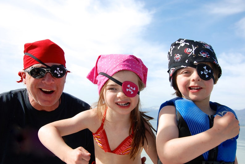 May include: Three individuals wearing pirate-themed accessories including a red headscarf, a pink polka-dot headscarf, and a black skull-and-crossbones headscarf. Each person wears a black eye patch featuring a white skull-and-crossbones graphic.