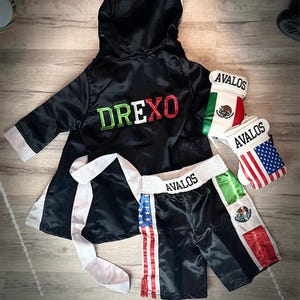 Op de afbeelding: Een zwarte boksset met een badjas, shorts en handschoenen. De badjas heeft de naam "DREXO" geborduurd in groen en rood. De shorts en handschoenen tonen de naam "AVALOS" en de vlaggen van de Verenigde Staten en Mexico.
