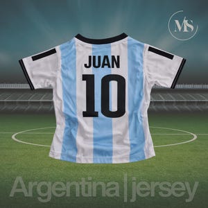 May include: Argentina jersey with the name "JUAN" and the number "10" on the back. The jersey has light blue and white vertical stripes, with black accents on the sleeves and collar. The text "Argentina jersey" is at the bottom.