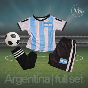 May include: A full soccer kit featuring a white and light blue striped jersey, black shorts with white stripes, and black socks with white stripes. A soccer ball and the text "Argentina | full set" are also visible.