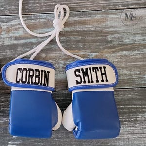 May include: Pair of blue and white boxing gloves with the names "CORBIN" and "SMITH" embroidered in black on the white wristbands. The gloves are connected by a white rope, suggesting they are decorative or for a small child.
