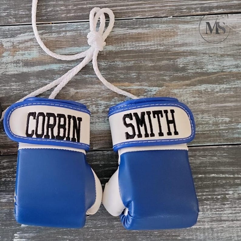May include: Pair of blue and white boxing gloves with the names "CORBIN" and "SMITH" embroidered in black on the white wristbands. The gloves are connected by a white rope, suggesting they are decorative or for a small child.