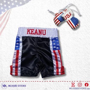 K&ouml;nnte beinhalten: Schwarze Satin-Boxershorts mit rot-wei&szlig;-blauen Streifen der amerikanischen Flagge. Die Shorts haben einen wei&szlig;en Bund mit dem Namen "KEANU" in rot gestickt.