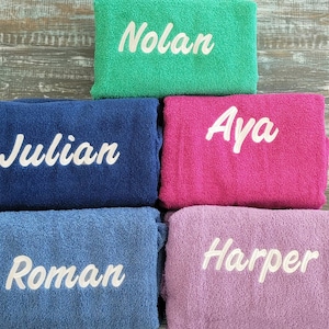 May include: Five towels with different names embroidered on them. The towels are stacked on top of each other. The names are Nolan, Aya, Julian, Roman, and Harper. The towels are in different colors: green, pink, blue, light blue, and purple.