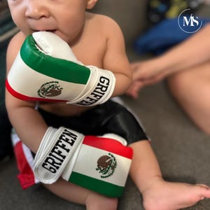 May include: White, red, and green boxing gloves with the word "GRIFFEN" in black lettering. The gloves feature the Mexican coat of arms. The gloves are on a baby.