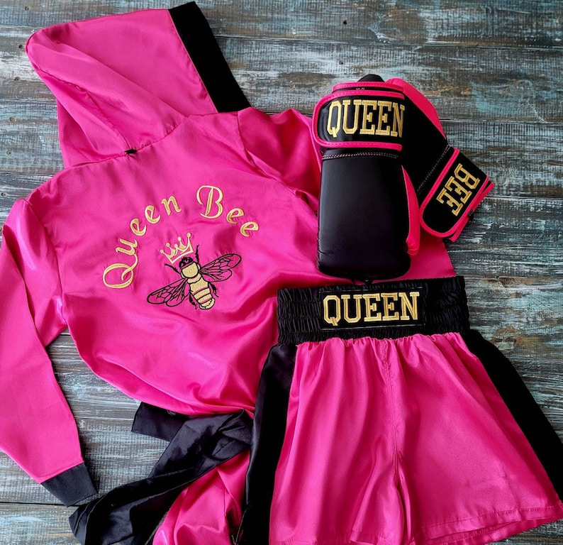 May include: A pink boxing robe, shorts, and gloves set. The robe has a black hood and trim, with "Queen Bee" and a bee embroidered in gold. The shorts have a black waistband with "QUEEN" in gold. The gloves are black and pink with "QUEEN" and "BEE" in gold.