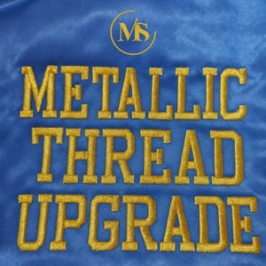 May include: A close-up of a blue fabric with gold embroidered text that reads "METALLIC THREAD UPGRADE". A gold circular logo with the letters "MS" is at the top.