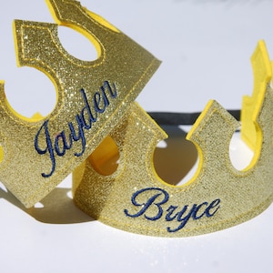 May include: Two gold glitter crowns with yellow felt trim. The crowns are personalized with the names "Jayden" and "Bryce" in blue embroidery.