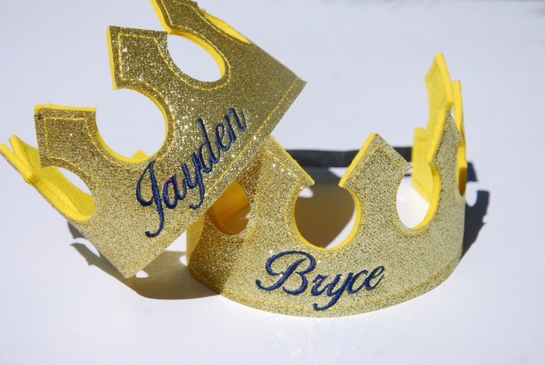 May include: Two gold glitter crowns with yellow felt trim. The crowns are personalized with the names "Jayden" and "Bryce" in blue embroidery.