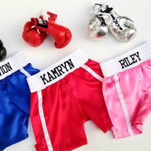 K&ouml;nnte beinhalten: Drei Paar personalisierte Boxershorts mit wei&szlig;em Besatz und passenden Boxhandschuhen. Die Shorts sind blau, rot und pink mit den Namen "Travivon", "Kamryn" und "Riley" auf dem Bund gestickt.