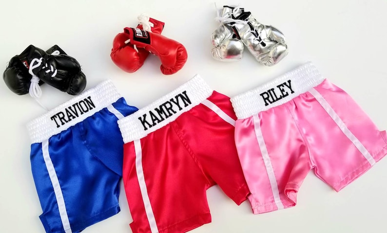 K&ouml;nnte beinhalten: Drei Paar personalisierte Boxershorts mit wei&szlig;em Besatz und passenden Boxhandschuhen. Die Shorts sind blau, rot und pink mit den Namen "Travivon", "Kamryn" und "Riley" auf dem Bund gestickt.