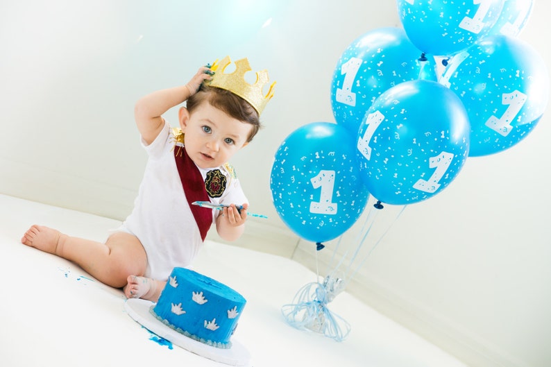 May include: A baby wearing a white shirt with a red sash and a gold crown, holding a plastic knife and looking at the camera. The baby is sitting in front of a blue and white cake with a number one on it. There are blue balloons with the number one on them in the background.