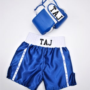 May include: Blue and white boxing shorts with white piping and the name "TAJ" embroidered on the waistband. A pair of blue and white boxing gloves with the name "TAJ" embroidered on the wrist strap.