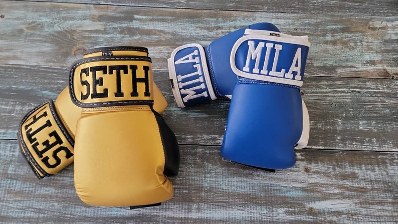 May include: Two boxing gloves, one pair in gold with "SETH" in black lettering, and another pair in blue with "MILA" in white lettering. The gloves are set against a weathered, blue-gray wooden surface.