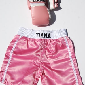May include: Pink satin boxing shorts with white trim and the name "Tiana" embroidered on the waistband. A matching pair of pink boxing gloves with white trim and the name "Tiana" on the wrist strap.