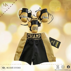 K&ouml;nnte beinhalten: Goldene Boxershorts mit schwarzem Besatz und dem gestickten Wort "CHAMP" am Bund. Drei goldene Mini-Boxhandschuhe mit den gestickten Worten "Lil Champ" und "Momma's gonna knock you out".