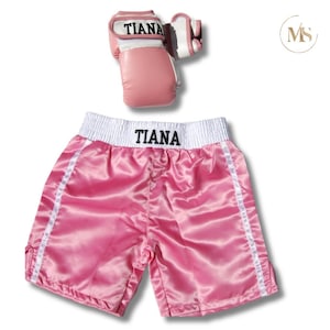 May include: Pink boxing gloves and shorts set. The gloves and shorts have the name "TIANA" printed on them. The shorts are a shiny pink satin with white trim and a white stripe down the sides. The gloves are pink with white accents.