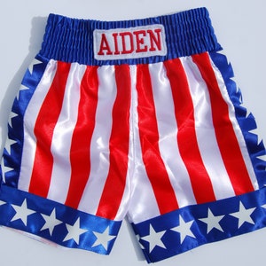 May include: Red, white, and blue satin boxing shorts with a blue waistband and white stars on the bottom. The shorts have the name "AIDEN" embroidered on the waistband.